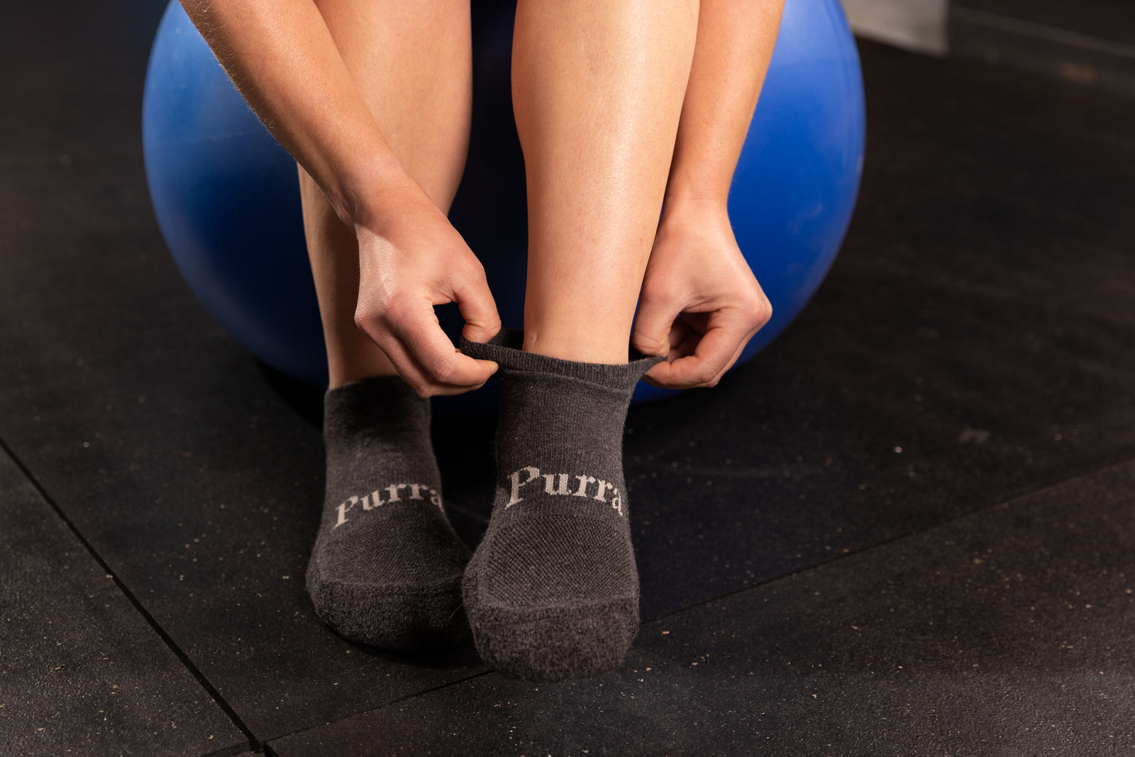 A woman sitting down wearing her bright blue exercise pants is preparing to exercise on the dark colored gym mat is seen adjusting her dark grey Purra No Show Socks.