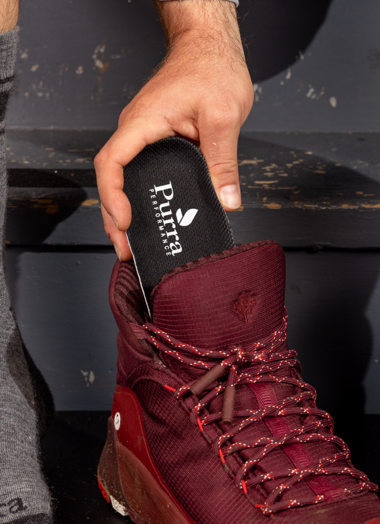 A man's hand is inserting a Purra insole into a dark red pair of hiking boots.  He is sittng on grey color wooden step to do this and is wearing the soon to be available Purra Merino Wool hiking socks