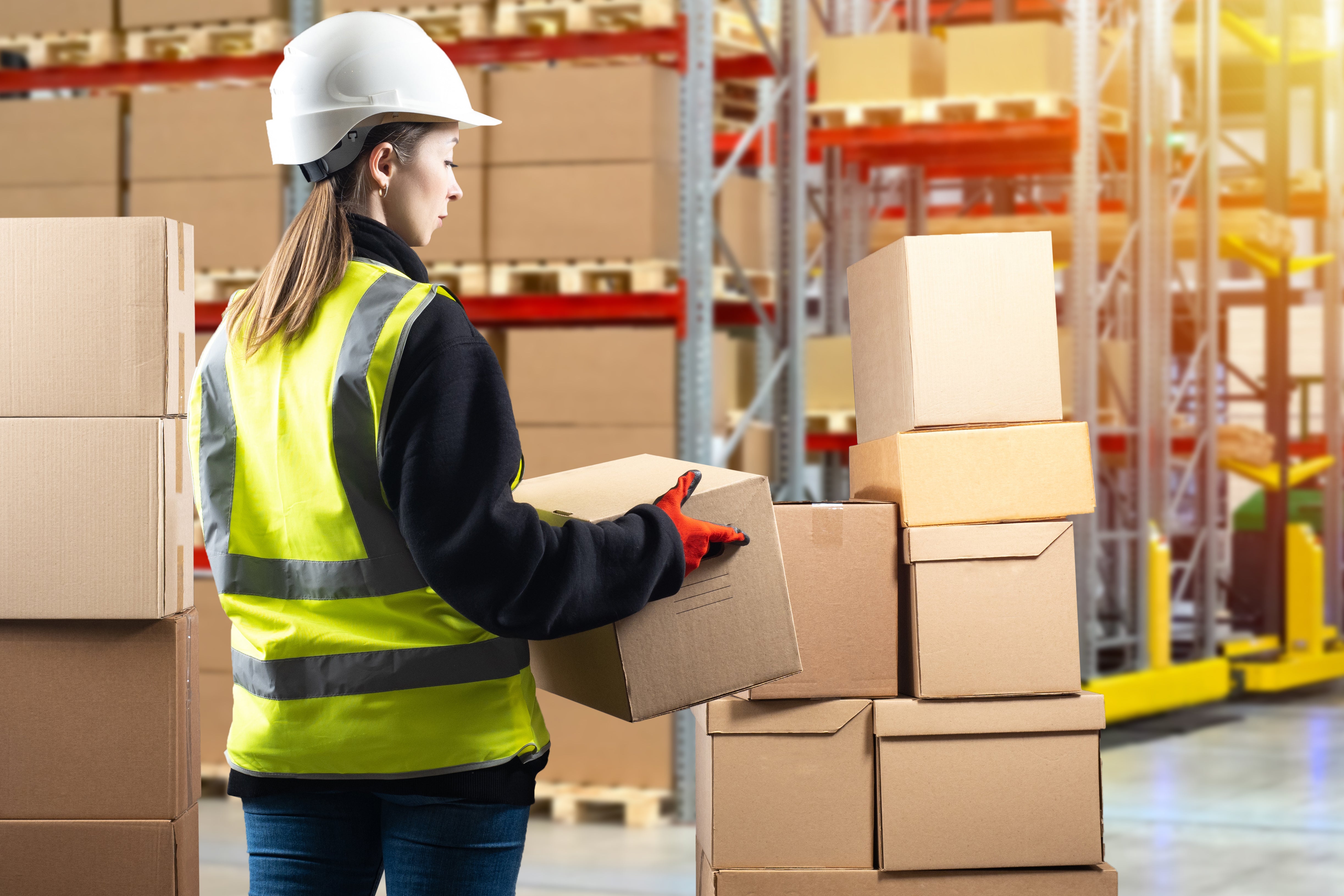 A warehouse working is shown in her hard hat and hi-viz vest stacking boxes.   This highlights the waste of boxes and cardboard shown on the racking unit behind her.