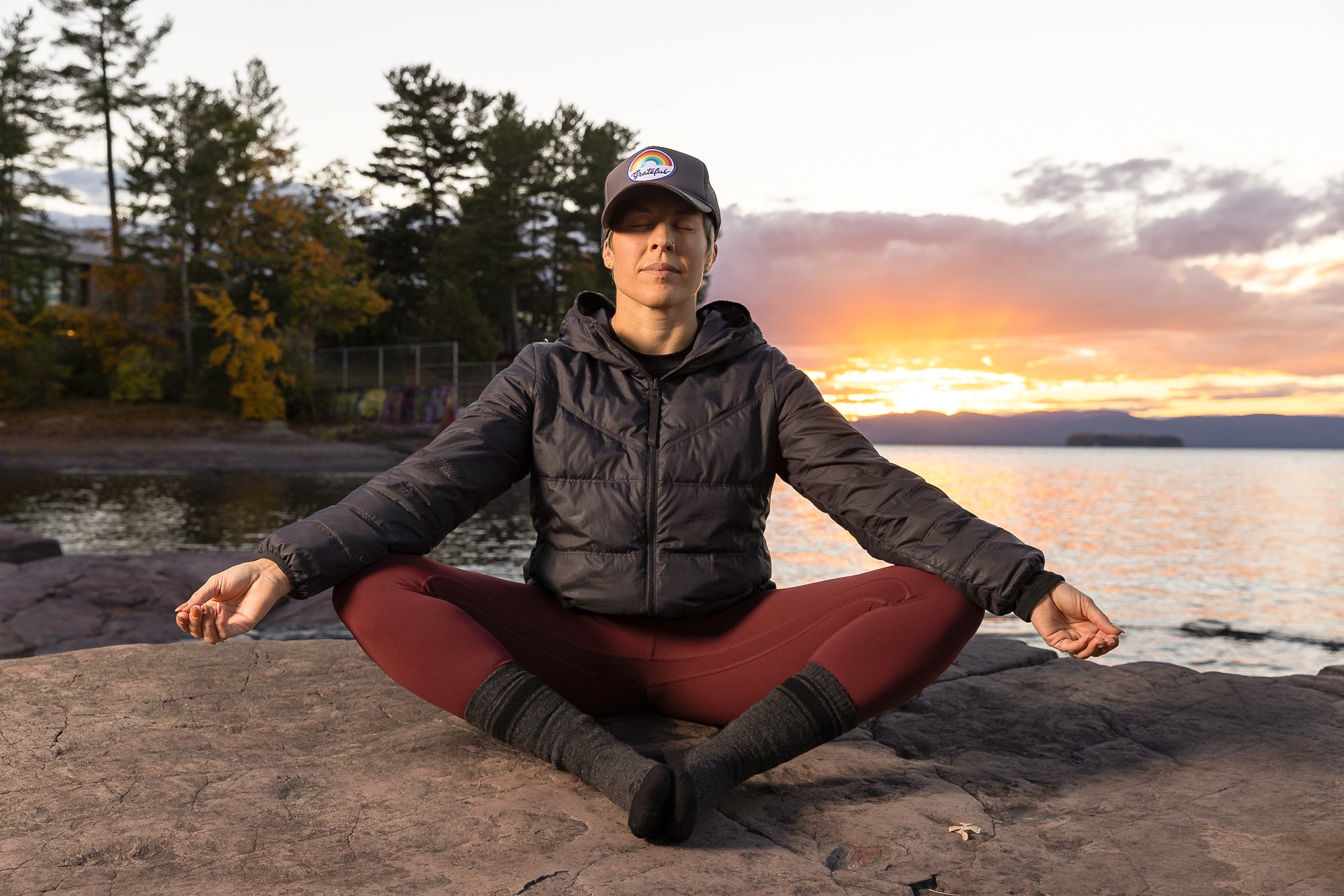 Woman sitting on a rock in a yoga position with her feet together meditating.  The sun is setting behind her reflected in the water.  She is wearing the dark grey Purra Crew Socks on her feet as well as dark colored leggings, an insulator jacket and a baseball cap