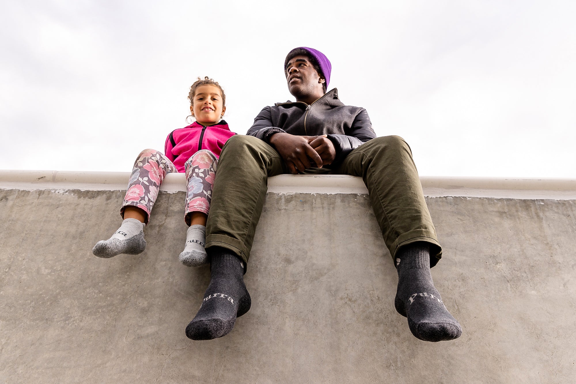 A view looking up at a dark skin toned father and his daughter sitting on a concrete wall.  The little girl is wearing pink flowery pants and bubblegum pink jacket.  On her feet she has the light grey Purra no show socks.  The father has khaki green long pants and a brown jacket with a purple beanie on his head.  On his feet he has the dark grey Purra Crew socks