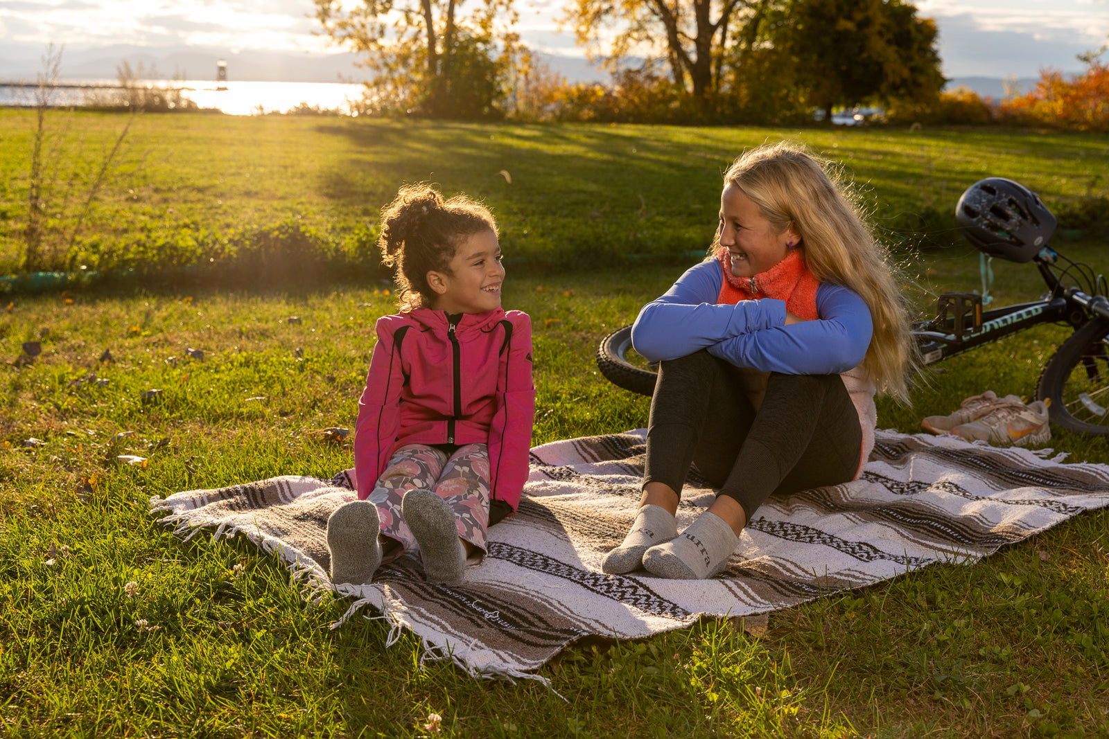 Two young girls are stopped after a bike ride sitting on a blanked with the lake shown behind them.  The younger girl on the left is wearing her bright pink jacket and floral pants with her light grey Purra No Show Socks and the older girl on the right with the bicycle behind her in her bright orange and blue jacket and dark colored pants is also wearing her No Show light grey socks.