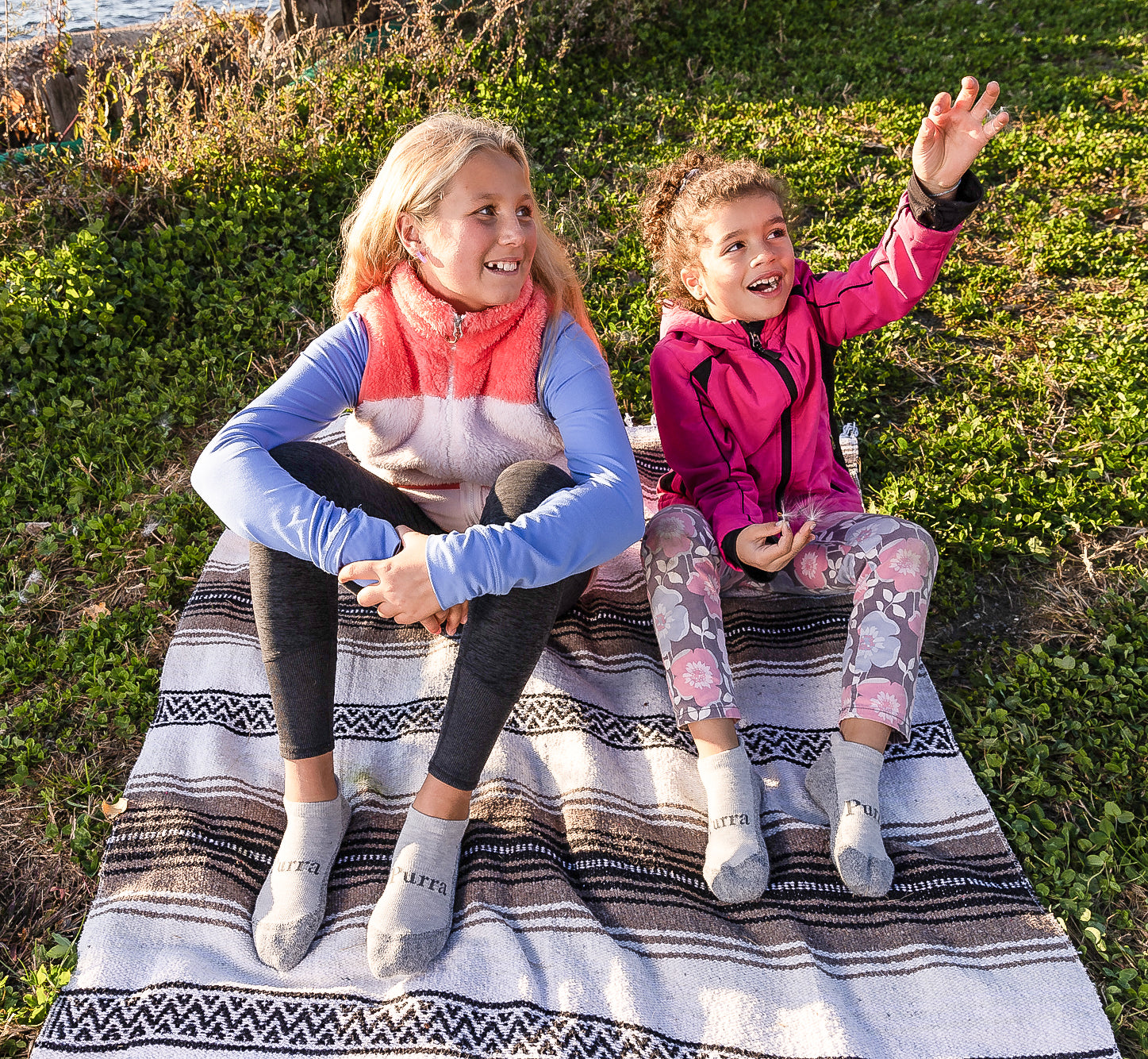 Two young girls are setting by the edge of a body of water on a blanket covering the grass.  The youngest on the right is wearing a bright pink jacket and floral leggings with her light grey Purra No Show Socks.  The older girl on the left is wearing a furry vest and black leggings also with her light grey Purra No Show socks