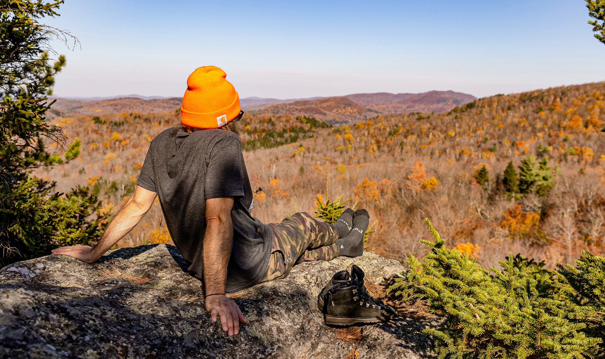 Landscape shot of fall colors in the background with a man sitting on a rock looking at the view.  He has his hiking boots on to his right and is wearing a pair of the Purra Comfort Crew socks.  He has on a bright orange beanie, grey t-shirt and camo colored pants.  Taking a rest during his hike up into the mountains.