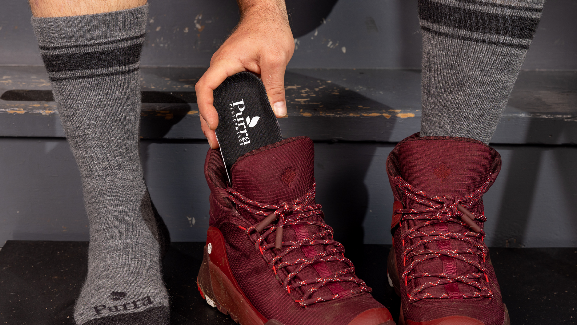 A man's hand is inserting a Purra insole into a dark red pair of hiking boots.  He is sittng on grey color wooden step to do this and is wearing the Purra Padded Boot Socks which will be available in the fall!